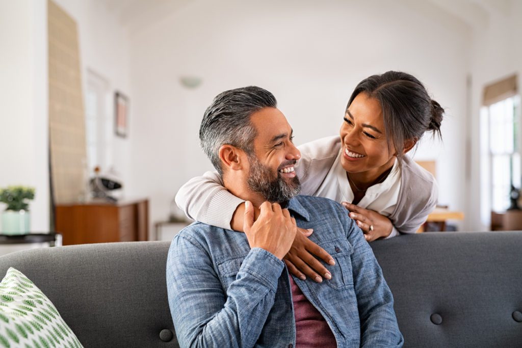 Smiling ethnic woman hugging her husband on the couch from behind in the living room.